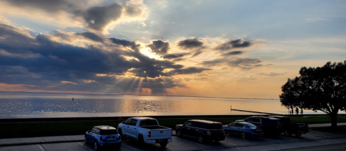 Sunset over a calm lake, with rays of light breaking through clouds and parked cars in the foreground.