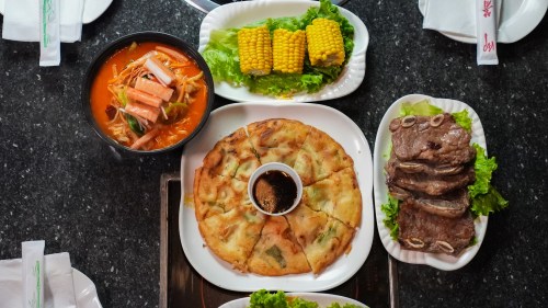 A table spread featuring a bowl of soup, corn on the cob, a pancake, and sliced meat on lettuce.