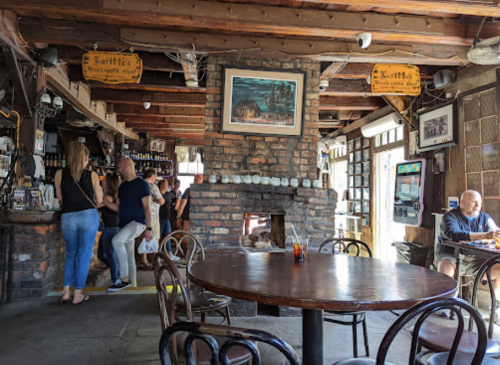 A cozy pub interior with wooden beams, a brick fireplace, and patrons enjoying drinks at tables.