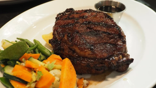 A grilled steak on a plate, accompanied by a side of mixed vegetables and a small cup of sauce.