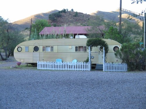 A vintage trailer home surrounded by greenery and a white picket fence, set against a mountainous backdrop.
