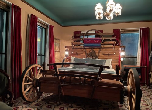 A rustic bedroom featuring a bed made from a wooden wagon, with red curtains and a chandelier overhead.
