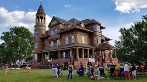 A large historic house with a tower, surrounded by people sitting on the lawn during an outdoor event.