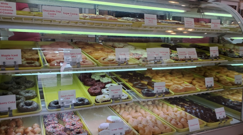 A display case filled with various donuts and cakes, labeled with names like "Chocolate Chip Cookie" and "Maple Iced Cake."