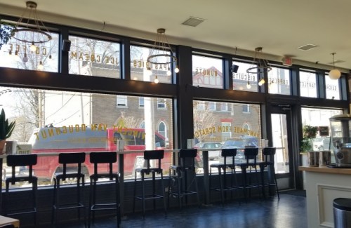 Interior of a café with tall stools by large windows, showcasing a view of the street outside. Bright and inviting atmosphere.