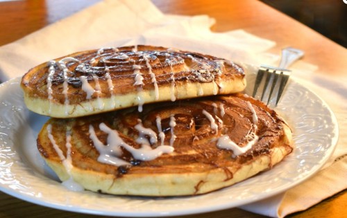 Two cinnamon roll pancakes drizzled with icing on a white plate, set on a wooden table with a fork beside them.