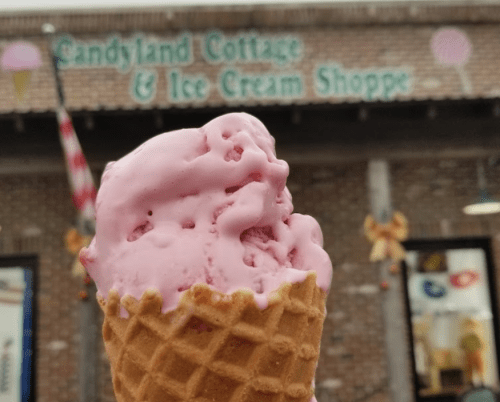 A close-up of a pink ice cream scoop in a waffle cone, with a shop sign reading "Candyland Cottage & Ice Cream Shoppe" in the background.