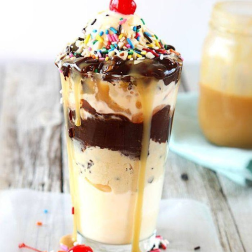 A tall glass filled with layers of ice cream, chocolate sauce, caramel, sprinkles, and topped with a cherry.