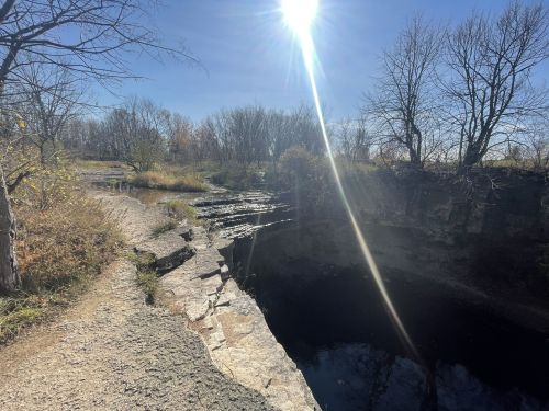 A sunlit landscape featuring a rocky path beside a deep, dark water-filled depression and bare trees in the background.