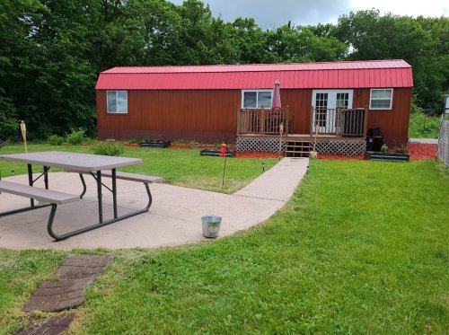 A wooden house with a red roof, surrounded by greenery, featuring a patio and picnic table in the foreground.
