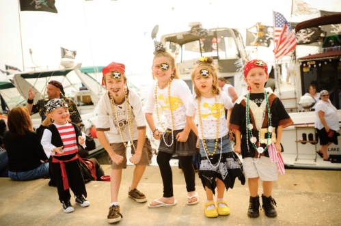 A group of children dressed as pirates, wearing bandanas and beads, posing happily at a festive waterfront event.