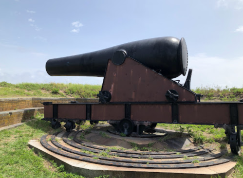 A large black cannon mounted on a rotating base, set against a clear blue sky and grassy landscape.