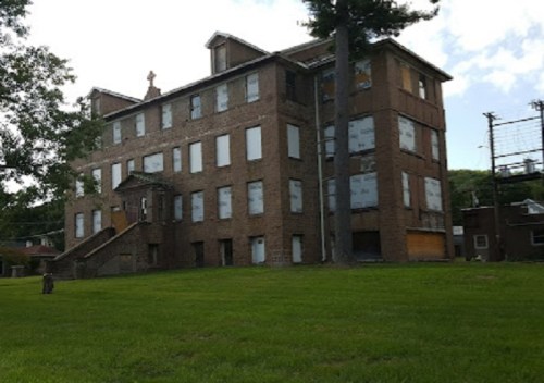 Abandoned brick building with boarded windows, set on a grassy area under a partly cloudy sky.