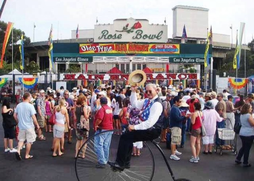 A crowd gathers at the Rose Bowl, with a man on a tall bicycle wearing a festive outfit and holding a hat.