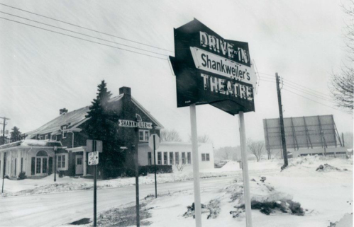 A snowy scene featuring the sign for Shankweiler's Drive-In Theatre, with a vintage building in the background.
