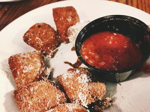 A plate of golden-brown fried snacks sprinkled with cheese, served with a small cup of marinara sauce.