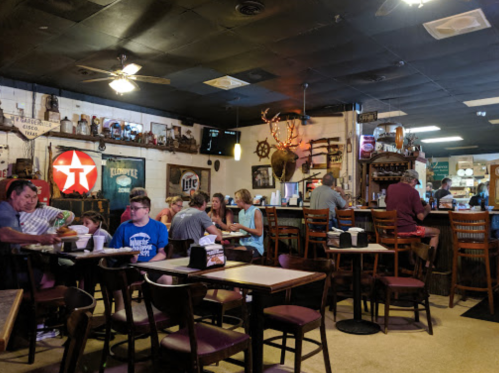 A cozy restaurant interior with patrons dining, a bar area, and vintage decor, including a neon sign and rustic elements.