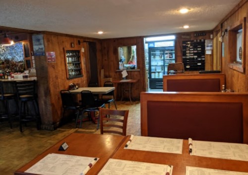 Cozy restaurant interior with wooden walls, tables set with menus, and a bar area in the background.