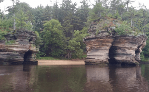 Two large rock formations rise from a calm river, surrounded by lush green trees and a sandy beach in the background.