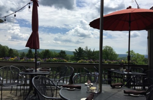 Outdoor dining area with black tables and chairs, red umbrellas, and a scenic view of trees and clouds in the background.