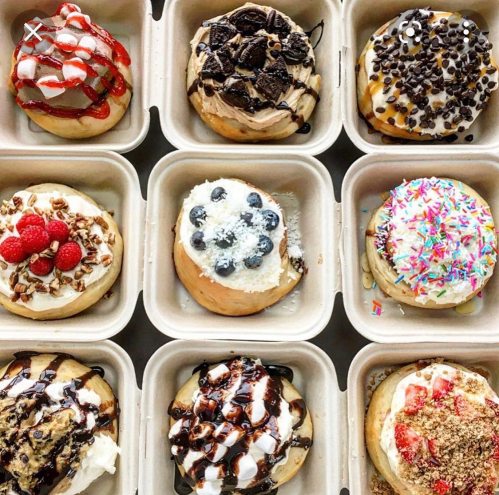 A variety of decorated cinnamon rolls topped with cream, fruits, and sprinkles in eco-friendly containers.