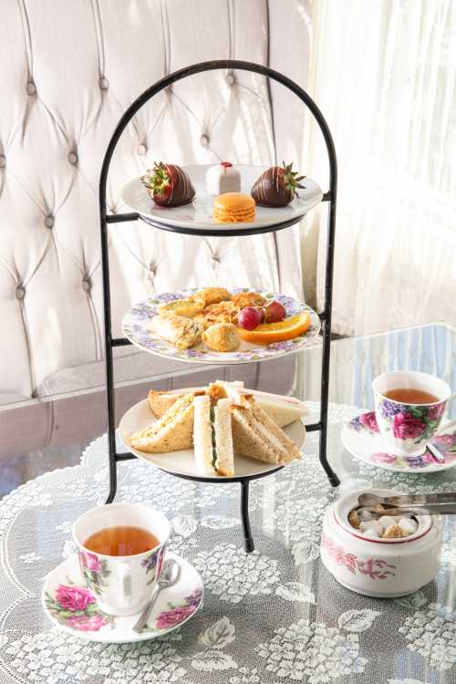 A tiered stand with assorted pastries, fruits, and sandwiches, accompanied by a cup of tea on a lace tablecloth.