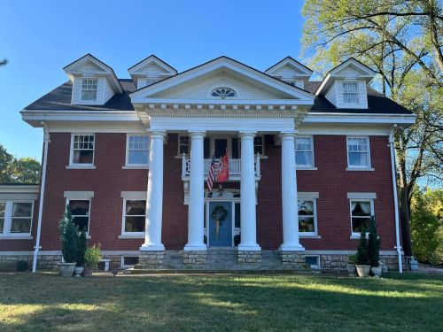 A large, elegant brick house with white columns, a front porch, and flags, surrounded by greenery under a clear blue sky.