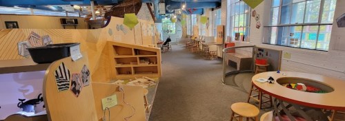 A bright, spacious play area with wooden structures, tables, and creative activity stations for children.