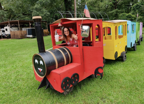 A woman gives a thumbs up from a colorful toy train in a grassy area, with trees and a camper in the background.
