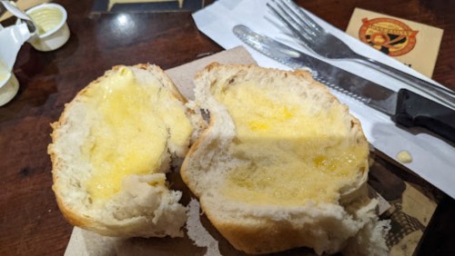 A split piece of bread with melted butter inside, placed on a brown paper napkin on a wooden table.