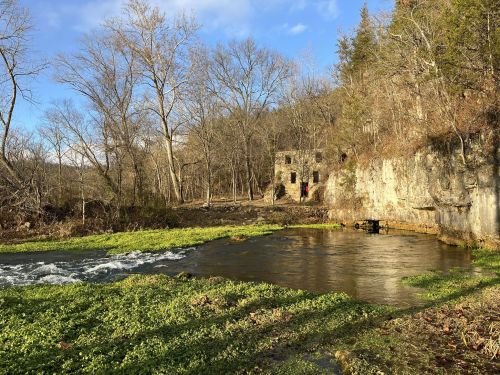 A serene river flows past a stone ruin, surrounded by trees and greenery under a clear blue sky.