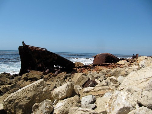 Rusty shipwreck on rocky shore with ocean waves and clear blue sky in the background.