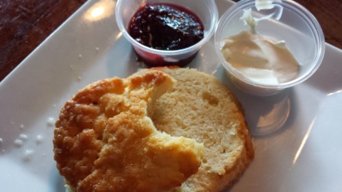 A plate with a biscuit, a bite taken out, and two small cups of jam and cream.