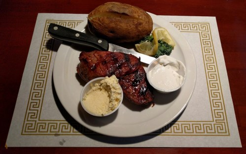 A plate with grilled steak, a baked potato, lemon, broccoli, and two sauces on the side.