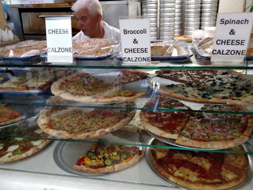 A display of various pizzas and calzones in a shop, with a person behind the counter. Signs indicate menu items.