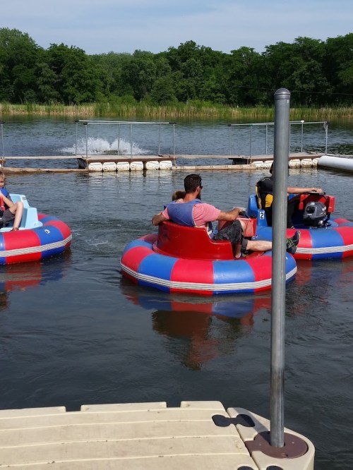 Two colorful bumper boats navigate a calm lake, surrounded by greenery and a wooden dock in the background.