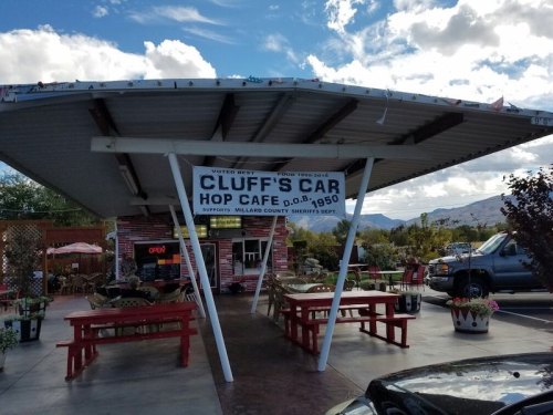 A roadside café with a large sign reading "Cluff's Car Hop Café," featuring outdoor seating and a scenic backdrop.
