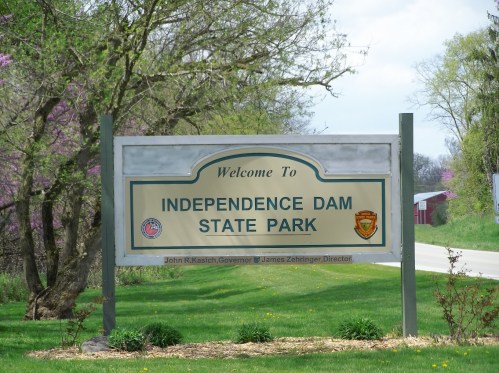 Welcome sign for Independence Dam State Park, featuring park logos and surrounded by greenery.