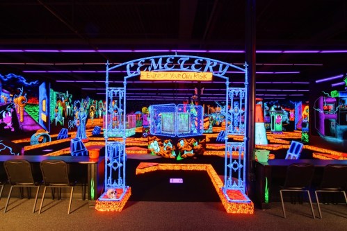 A vibrant, neon-lit mini-golf course featuring a spooky cemetery theme with glowing decorations and a sign reading "Enter at Your Own Risk."