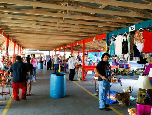 A bustling market scene with vendors and shoppers under a wooden structure, showcasing various goods and clothing.