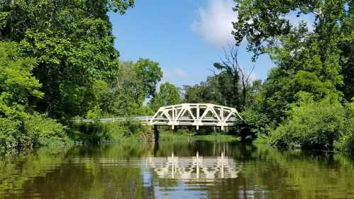 A white bridge spans a calm river, surrounded by lush green trees and a clear blue sky, reflecting in the water.