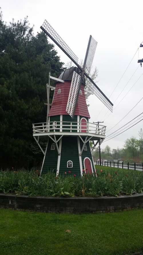 A colorful windmill surrounded by greenery and flowers, with power lines in the background on a cloudy day.