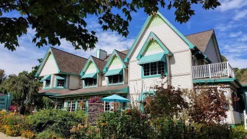 A charming, multi-gabled house with turquoise accents, surrounded by colorful flowers and greenery under a blue sky.