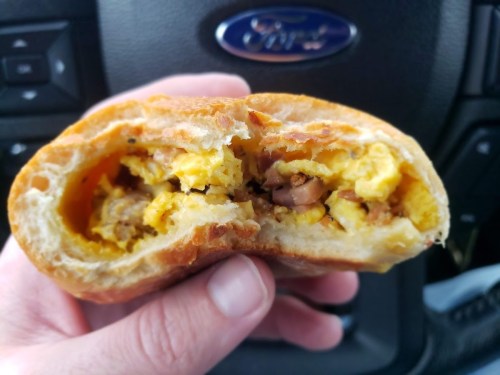 A hand holding a half-eaten breakfast sandwich with scrambled eggs and meat, set against a car interior background.