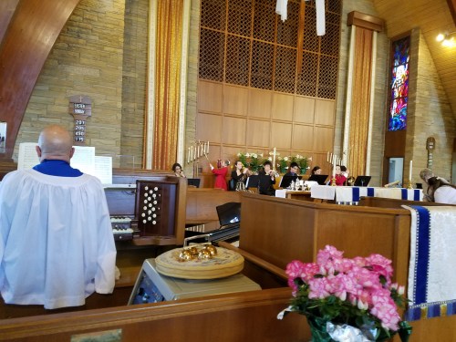 A church service in progress, featuring musicians and a congregation, with stained glass windows and floral arrangements.
