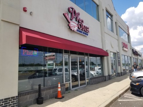 Exterior of Pho One restaurant with a red awning and neon sign, located in a shopping plaza on a sunny day.
