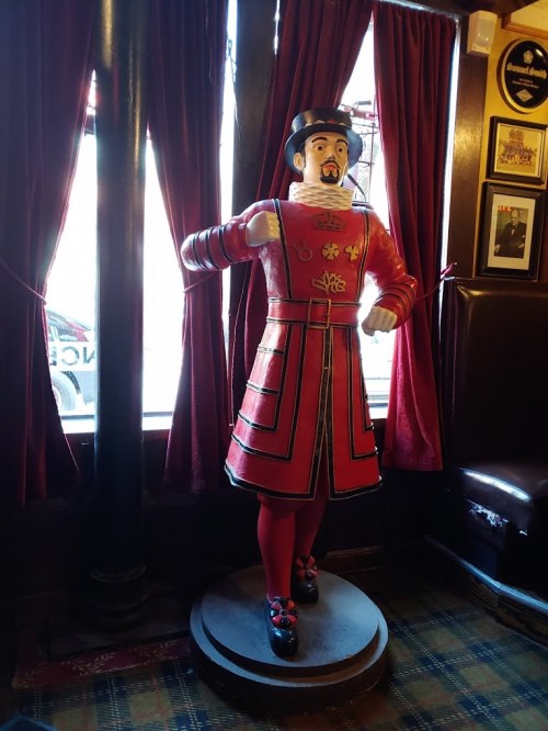 A colorful statue of a beefeater in a red uniform stands in a pub, surrounded by dark curtains and wooden decor.