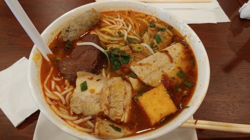 A bowl of spicy noodle soup with various meats, vegetables, and garnishes, served with chopsticks.