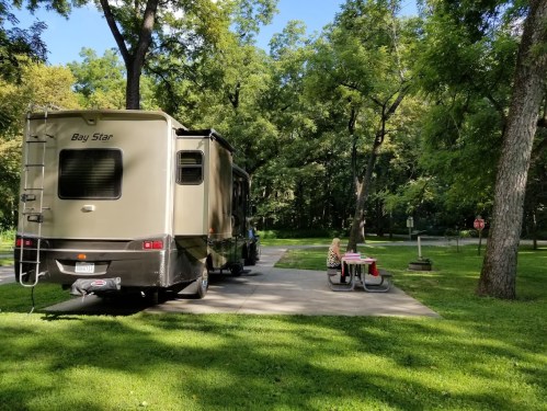 A parked RV in a green campsite, with a person sitting at a picnic table nearby under the trees.