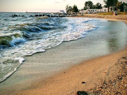 Waves gently lap at a sandy beach, with trailers in the background and a calm sea under a clear sky.